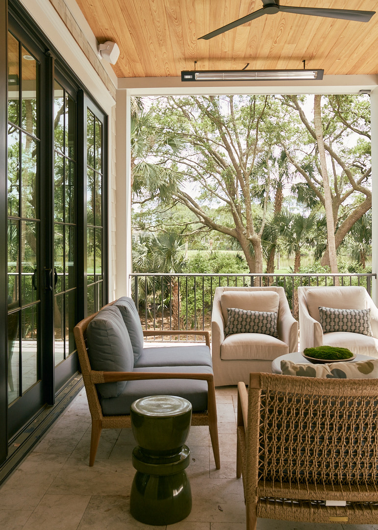 Covered outdoor living space with woven lounge seating, black steel French doors, tongue-and-groove wood ceiling, and tranquil views of mature live oaks and native Kiawah Island landscaping.