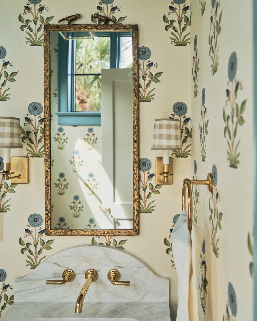 Charming powder room on Kiawah Island featuring floral botanical wallpaper, a custom marble vanity backsplash, unlacquered brass wall-mounted faucet, antique-style framed mirror, and tailored gingham shade sconces. Designed by PWD Studio, the space blends timeless craftsmanship, English cottage influences, and refined coastal elegance.