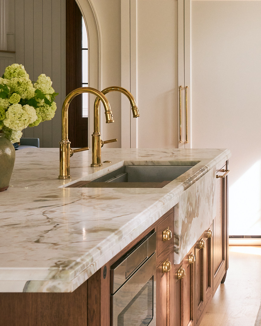 Furniture-style walnut kitchen island with waterfall marble countertop, integrated prep sink, brass faucets, and custom cabinetry in a refined coastal kitchen design.