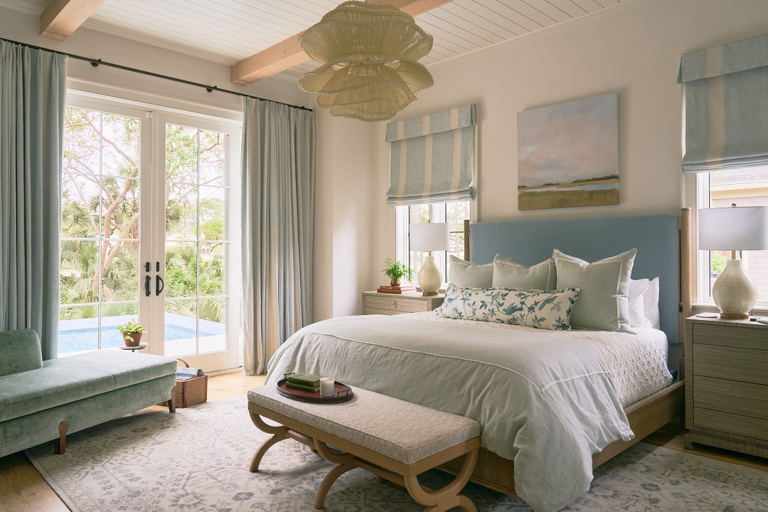 Serene primary bedroom featuring a sky-blue upholstered bed, custom drapery, woven statement chandelier, and French doors opening to a private pool terrace.
