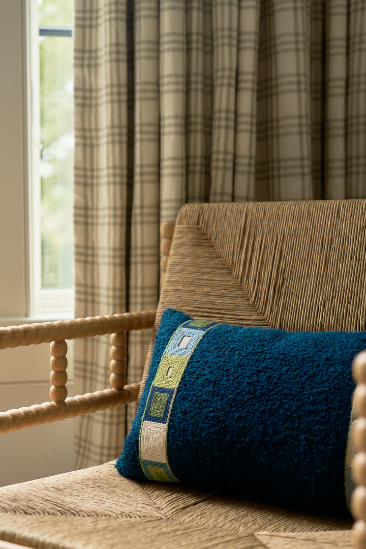 Woven accent chair styled with a vibrant blue embroidered pillow, layered textiles, and tailored plaid drapery in a relaxed coastal guest bedroom.