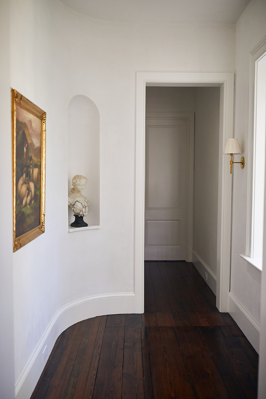A softly lit historic hallway with white plaster walls, curved baseboards, and rich heart pine flooring. A custom arched wall niche displays a carved marble bust on a stone shelf, while an antique gilt-framed landscape painting and polished brass wall sconce add timeless character. The restrained palette and architectural details create a refined, gallery-like transition space in this thoughtfully restored Charleston home.
