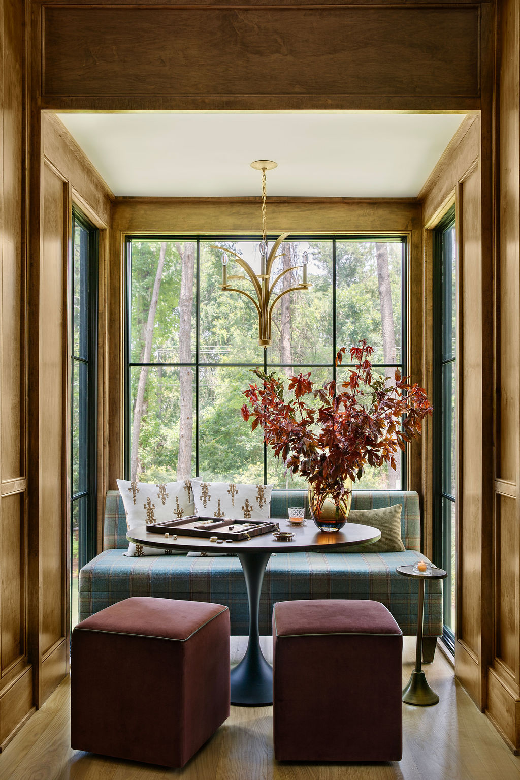 Luxury game room by PWD Studio featuring custom wood paneled walls, a plaid banquette, upholstered ottomans, a round game table, brass chandelier, and floor to ceiling black framed windows overlooking the landscape.