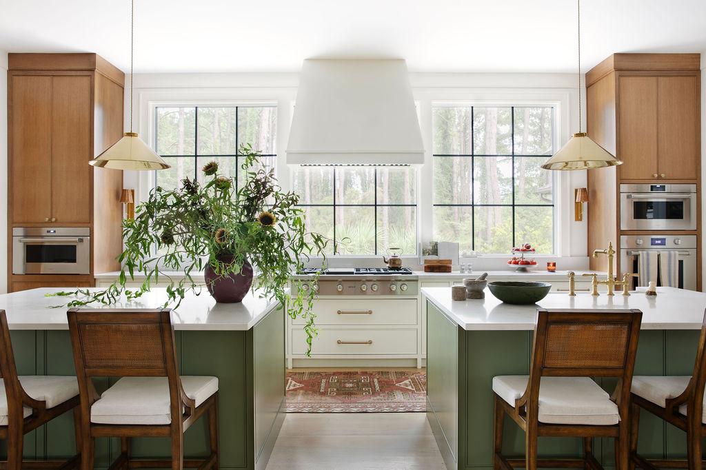 Luxury kitchen by PWD Studio featuring green island cabinetry, natural wood cabinets, white stone countertops, brass pendant lighting, a plaster range hood, and expansive black framed windows.