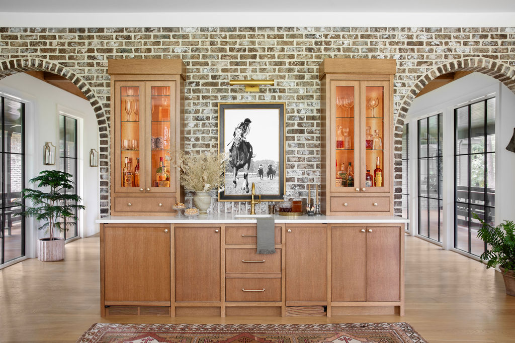Custom home bar by PWD Studio featuring natural wood cabinetry, glass front display cabinets, a white stone countertop, brass fixtures, antique brick, and arched openings framed by black steel windows.