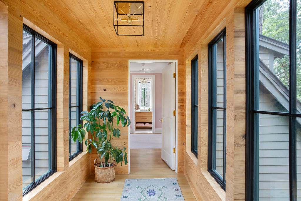 Light filled wood paneled hallway by PWD Studio featuring natural wood walls and ceiling, black framed windows, a geometric lantern, potted greenery, and views into a soft pink bedroom.