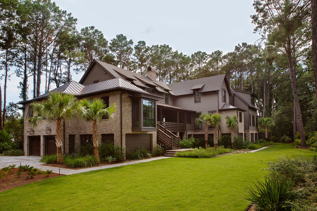 Lowcountry home exterior by PWD Studio featuring brick and wood siding, metal roof accents, black framed windows, palm trees, and lush landscaping surrounded by mature pines.