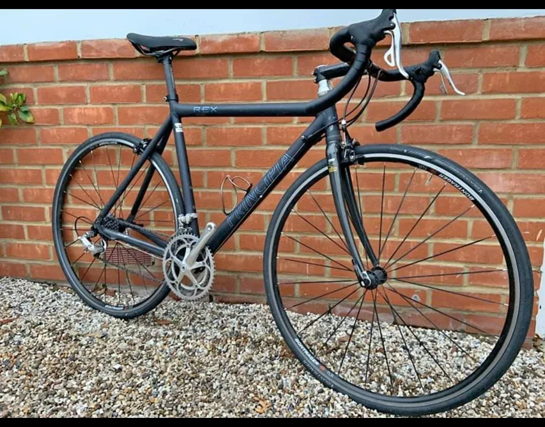 A black Principia Rex road bike in front of a red brick wall