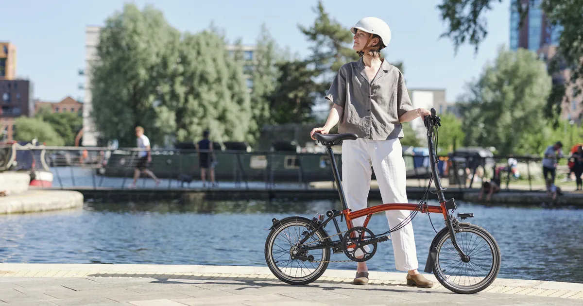 Orange Brompton folding bike with a steel frame, parked on the bank of an urban canal with a cyclist in the background