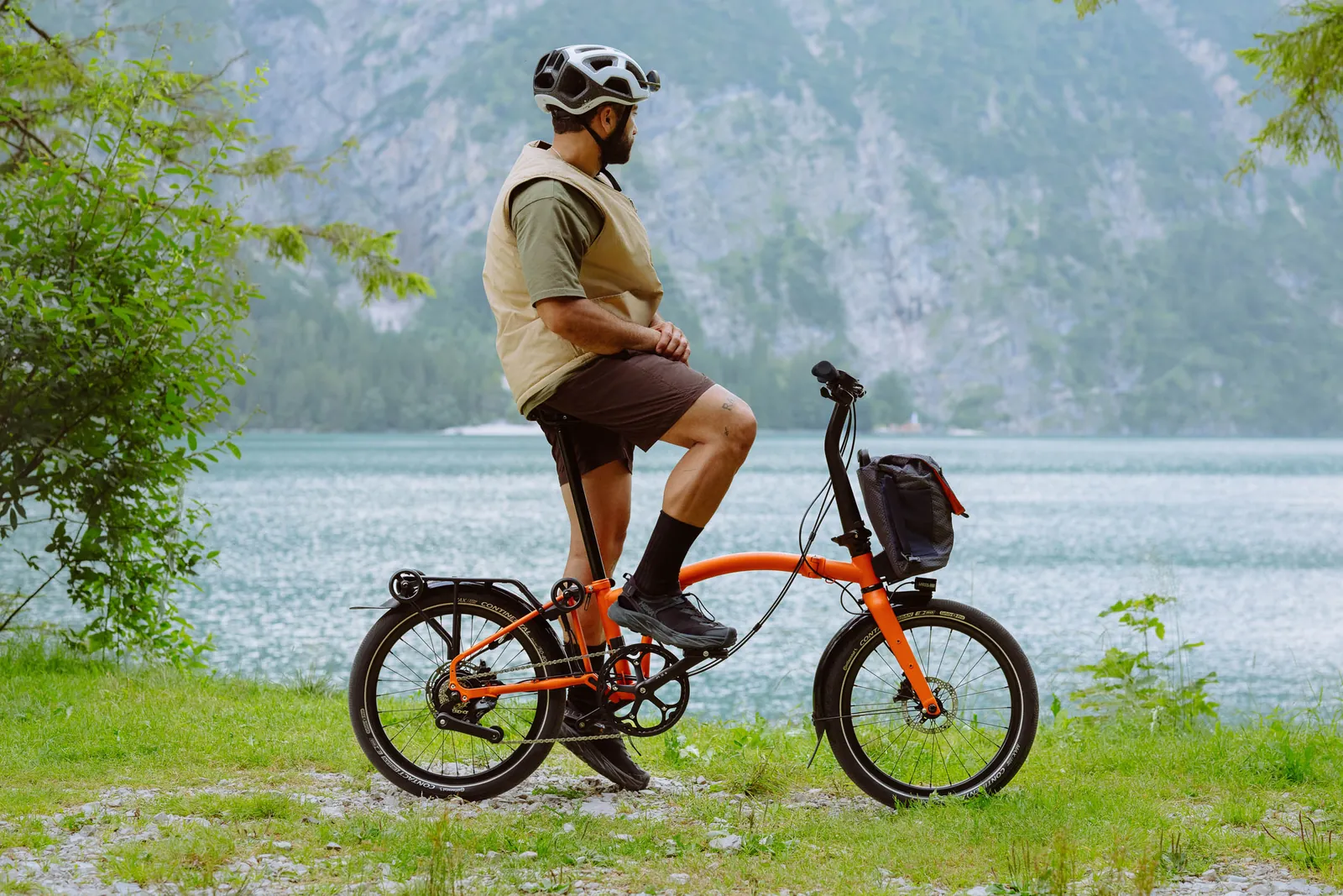 An orange Brompton folding bike by a lake, with a cyclist wearing a helmet and a beige vest