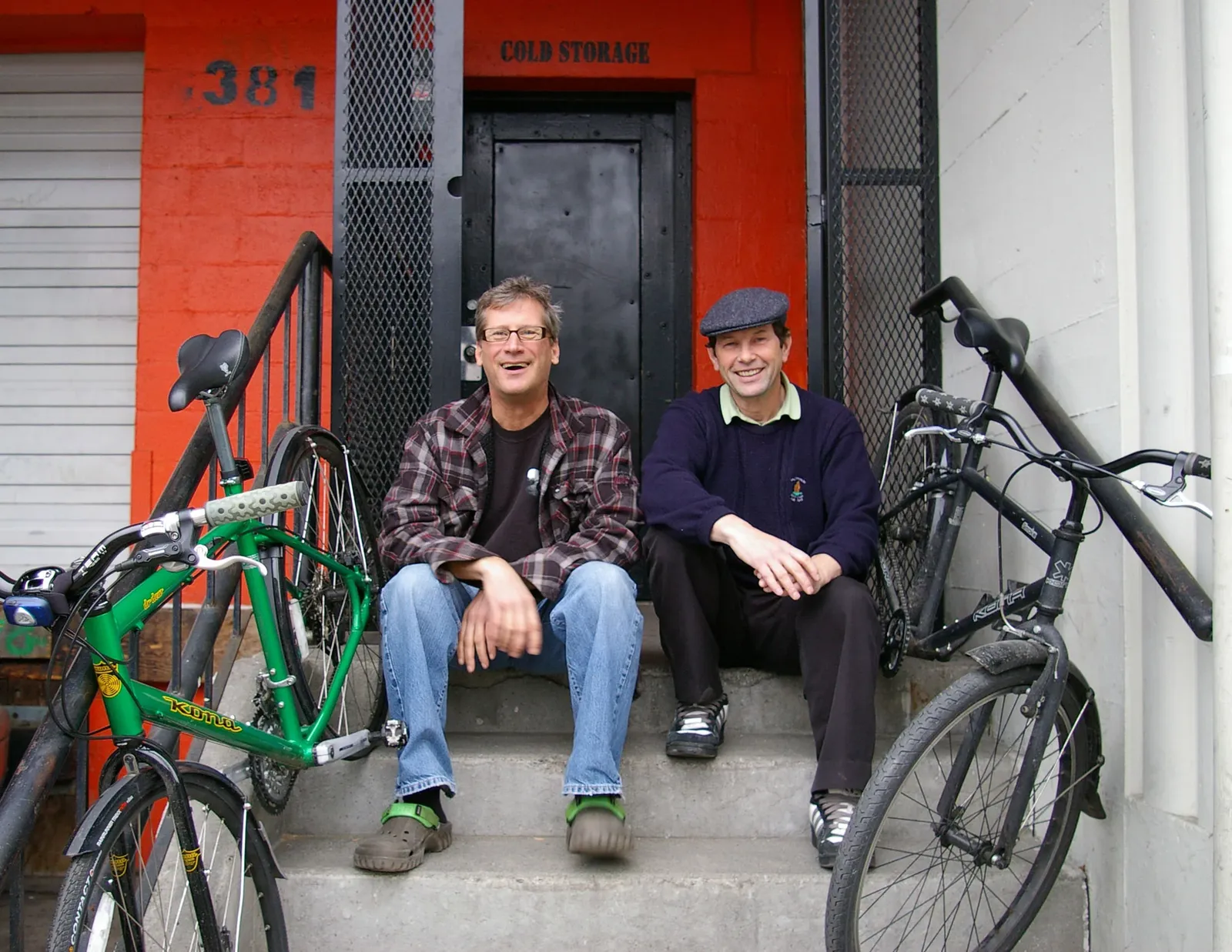 Two black and green Kona bikes leaning against concrete steps in front of an orange wall