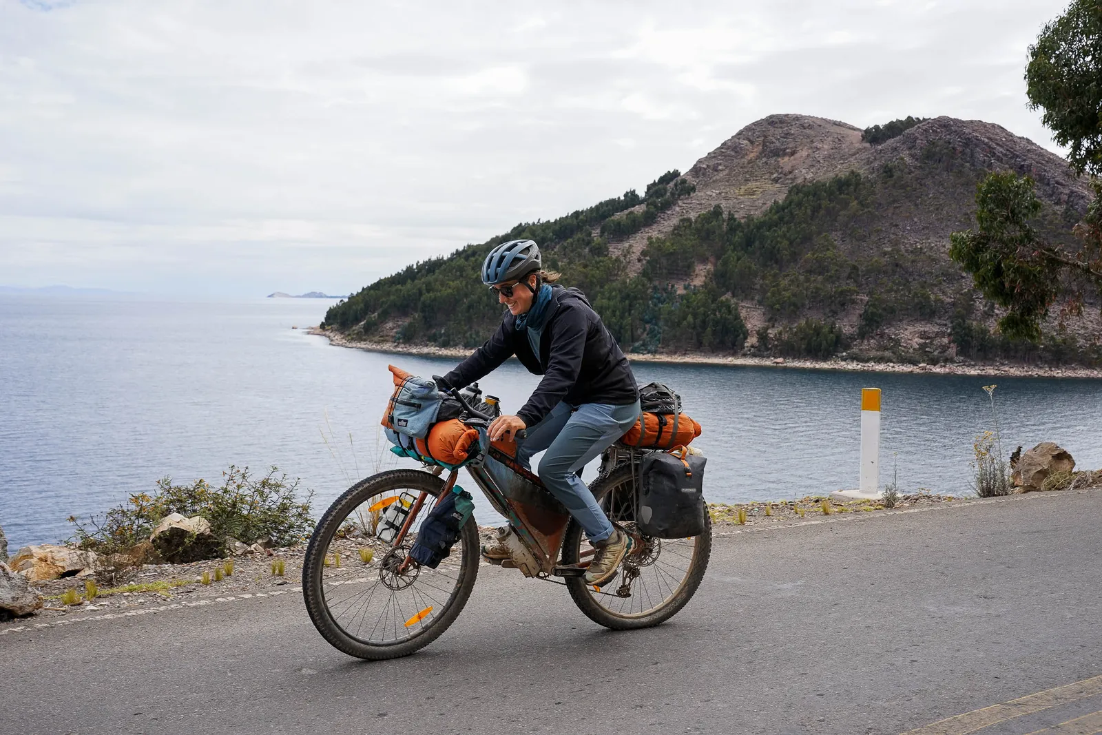 Kona bike on a coastal road, sturdy frame, orange panniers, fully equipped cyclist, mountainous landscape in the background