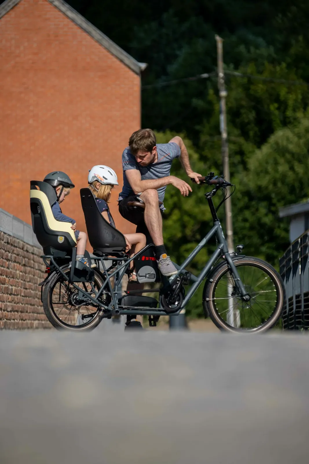 Gray Bike 43 cargo bike with child seats, sturdy frame, cobblestone street, red brick wall in the background