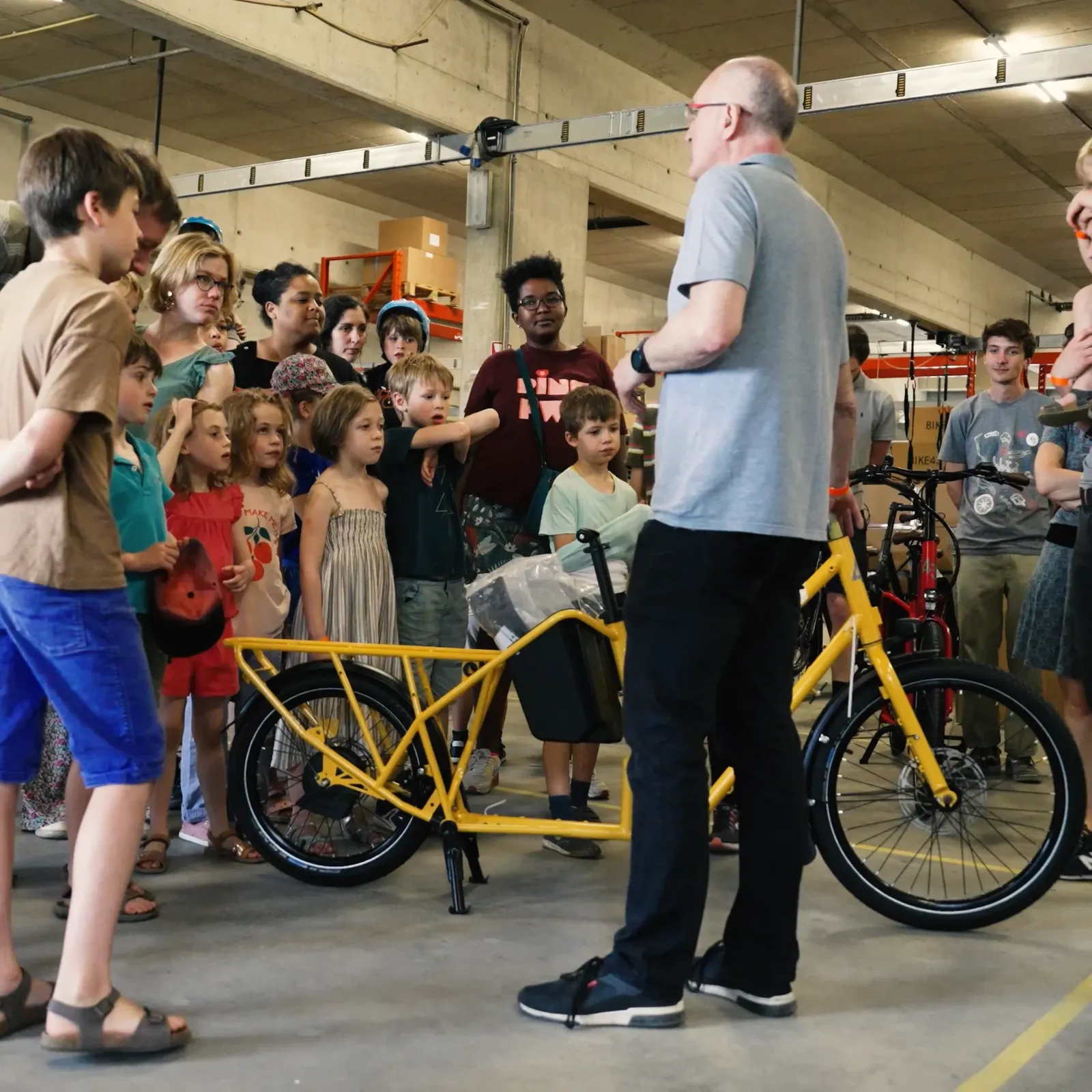 A yellow Bike 43 cargo bike indoors, with a sturdy frame, surrounded by people