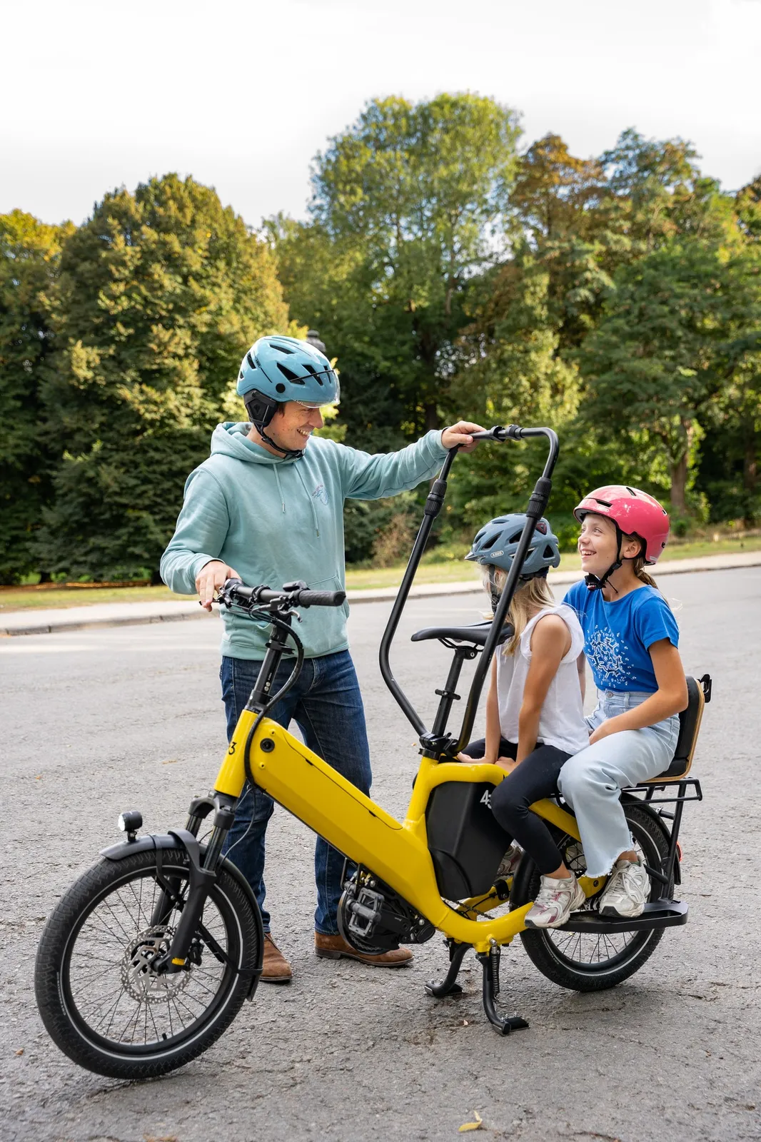 A yellow Bike 43 cargo bike with two children seated, an adult riding alongside, and a park in the background