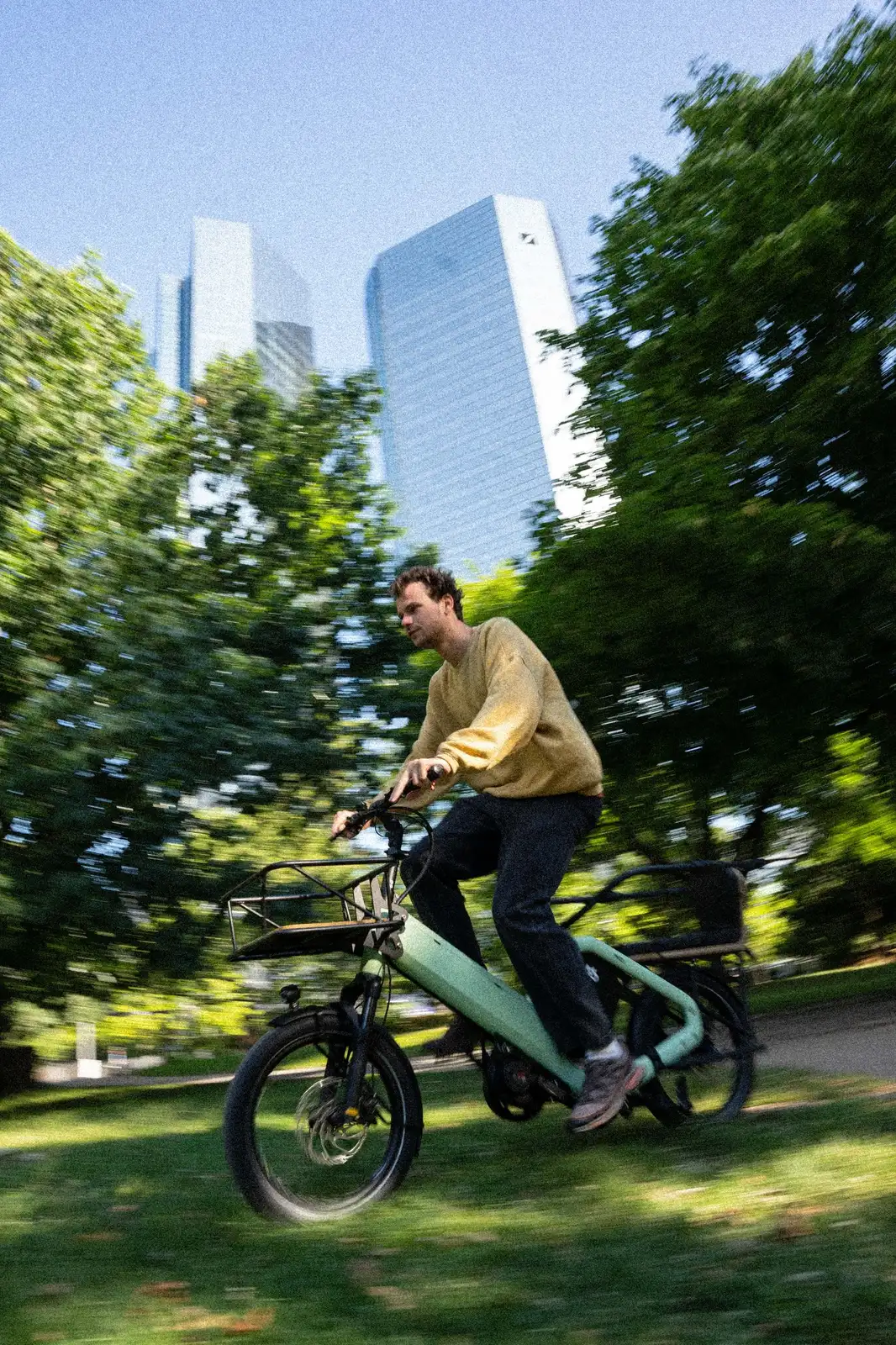 A green Bike 43 cargo bike riding through an urban park with skyscrapers in the background