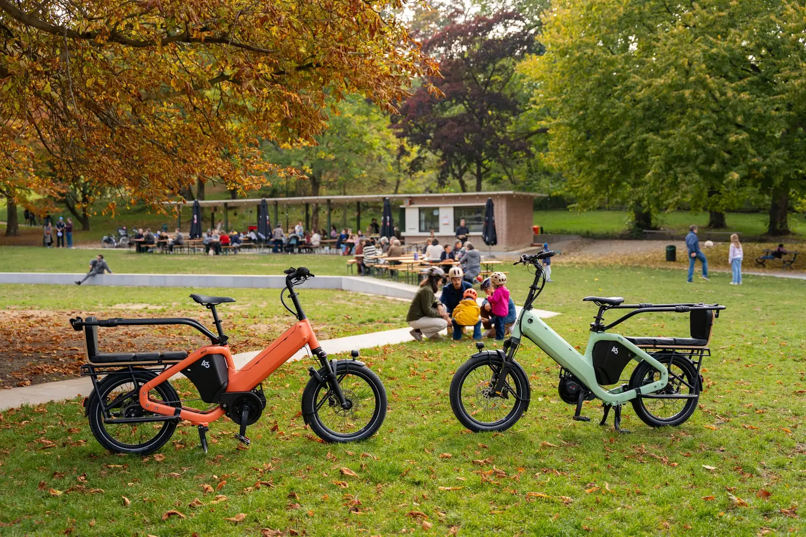 Two orange and green Bike 43 electric cargo bikes on the lawn with families in the background