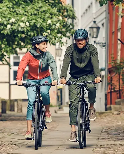 Two people riding black VSF Fahrradmanufaktur bicycles on a paved path in town, with trees in the background