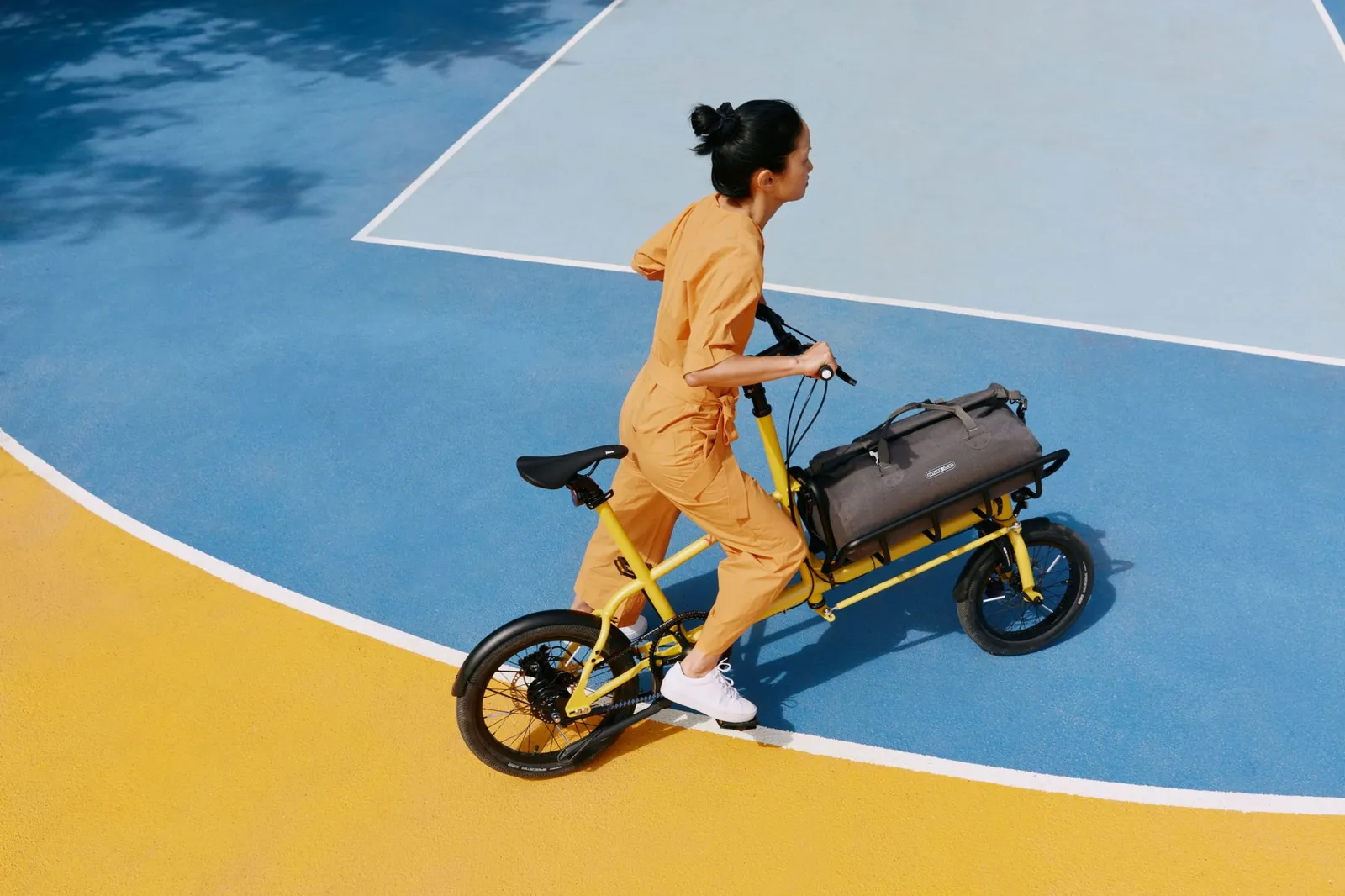 A yellow Yoonit cargo bike on a multicolored background, with a woman in an orange jumpsuit pedaling