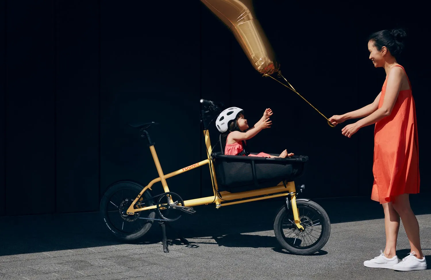 Yoonit yellow cargo bike with a child in the front seat and an adult in a red dress
