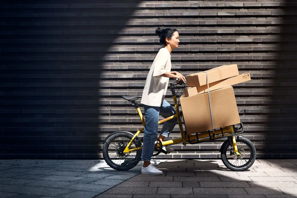 A yellow Yoonit cargo bike loaded with boxes in front of a dark brick wall