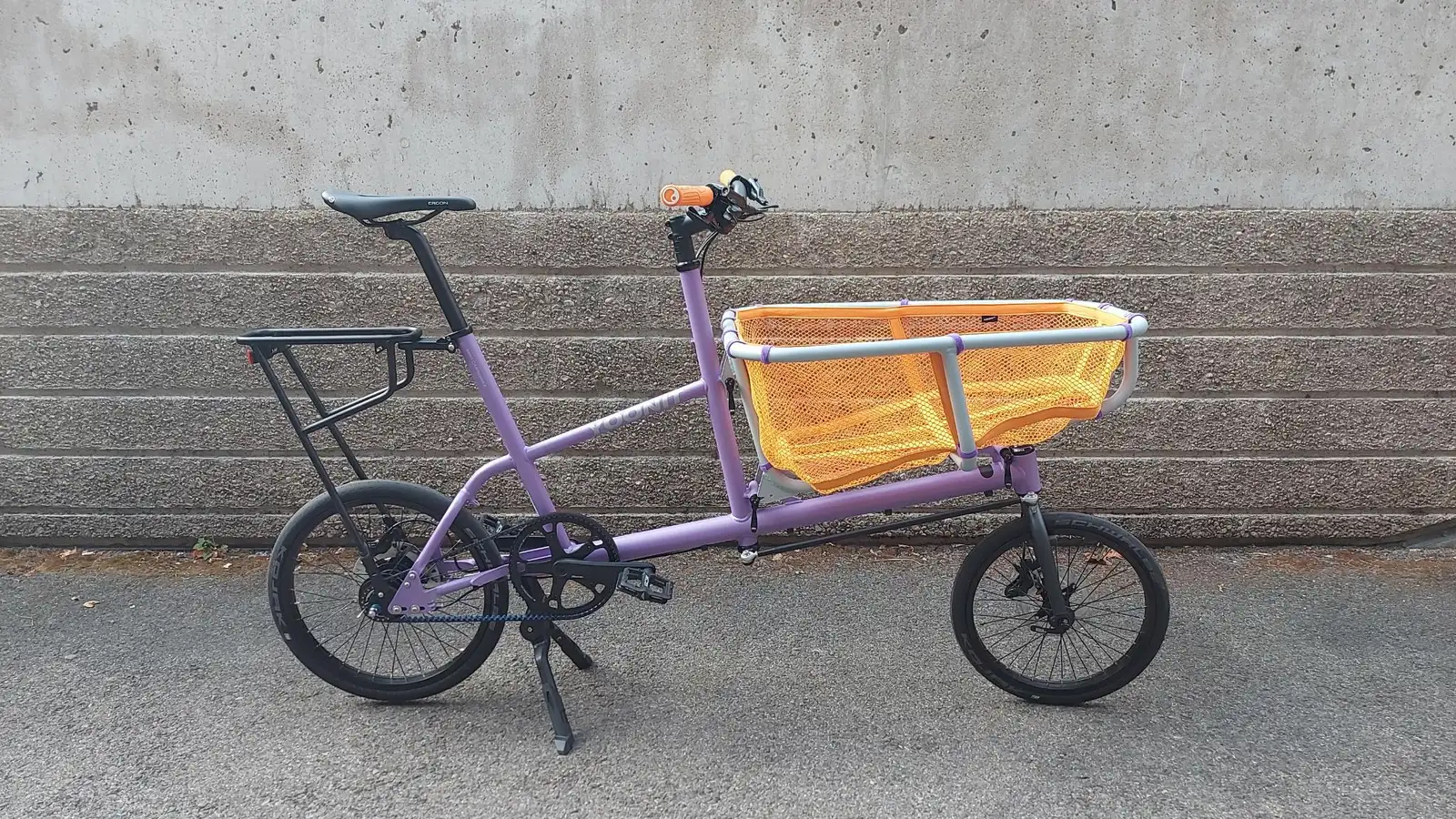 Purple Yoonit cargo bike with an orange basket, compact frame in front of a concrete wall