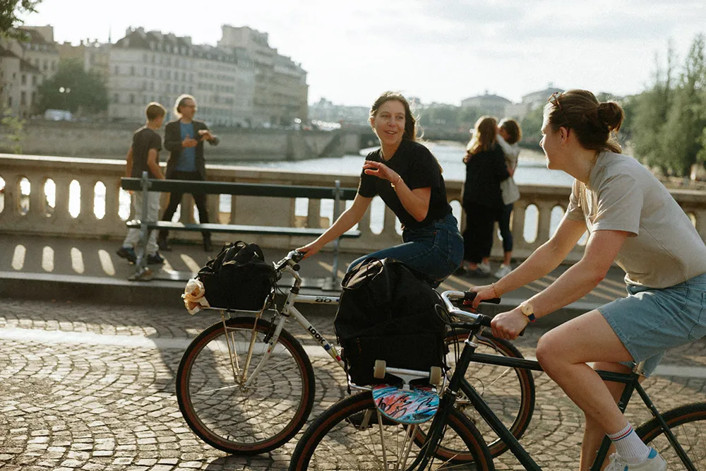 Two gray Pelago bikes with baskets, smiling cyclists in the city, with a bridge in the background