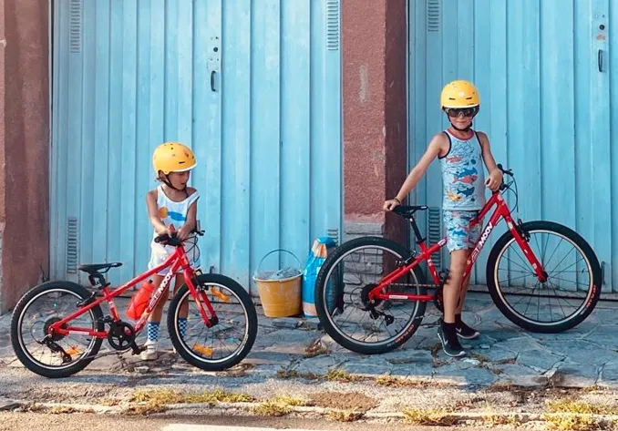 Two children wearing yellow helmets with red BeMoov bikes in front of blue doors