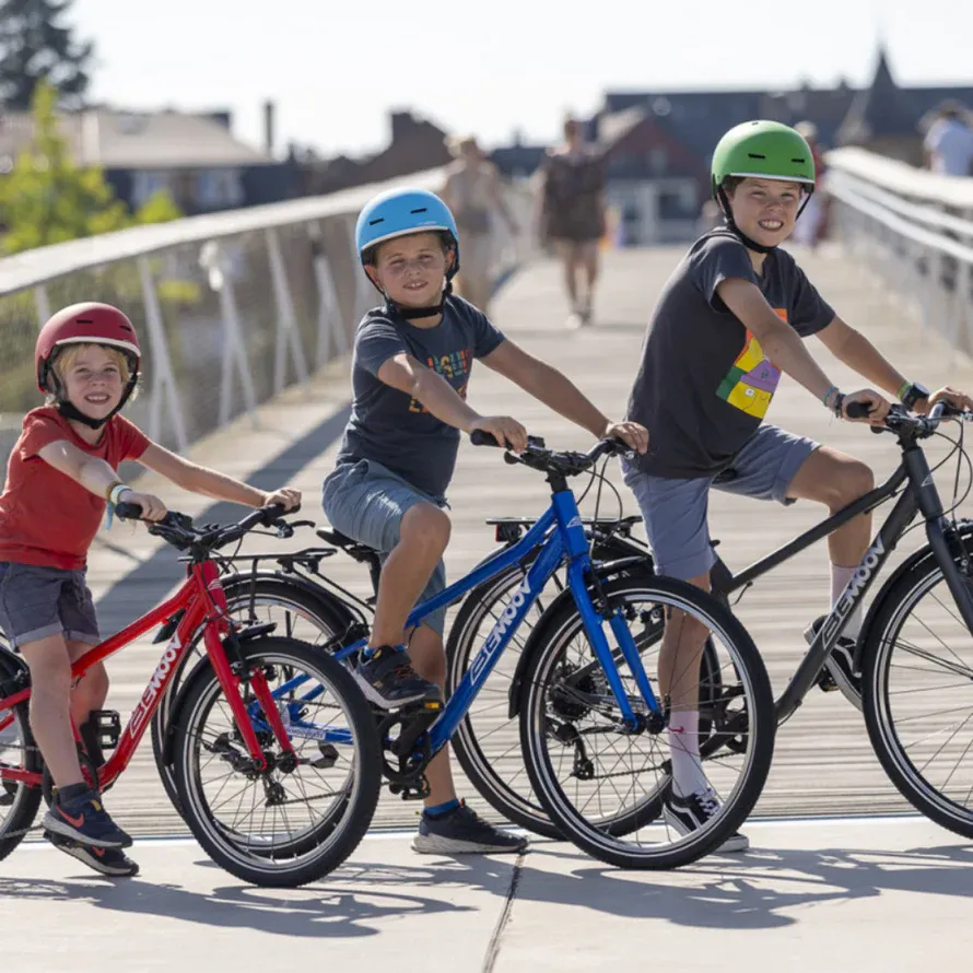 Red, blue, and black BeMoov bikes on a bridge with children wearing colorful helmets