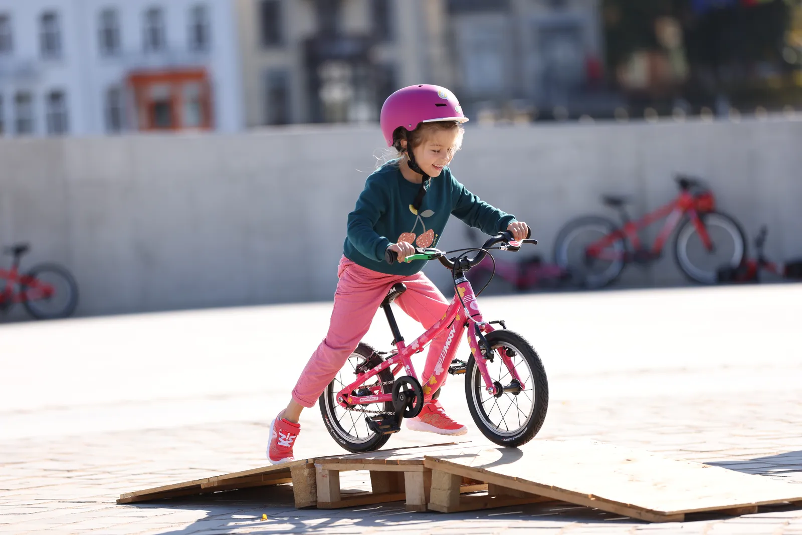 Child on a pink BeMoov bike, wearing a pink helmet, with a lightweight frame and an urban setting in the background
