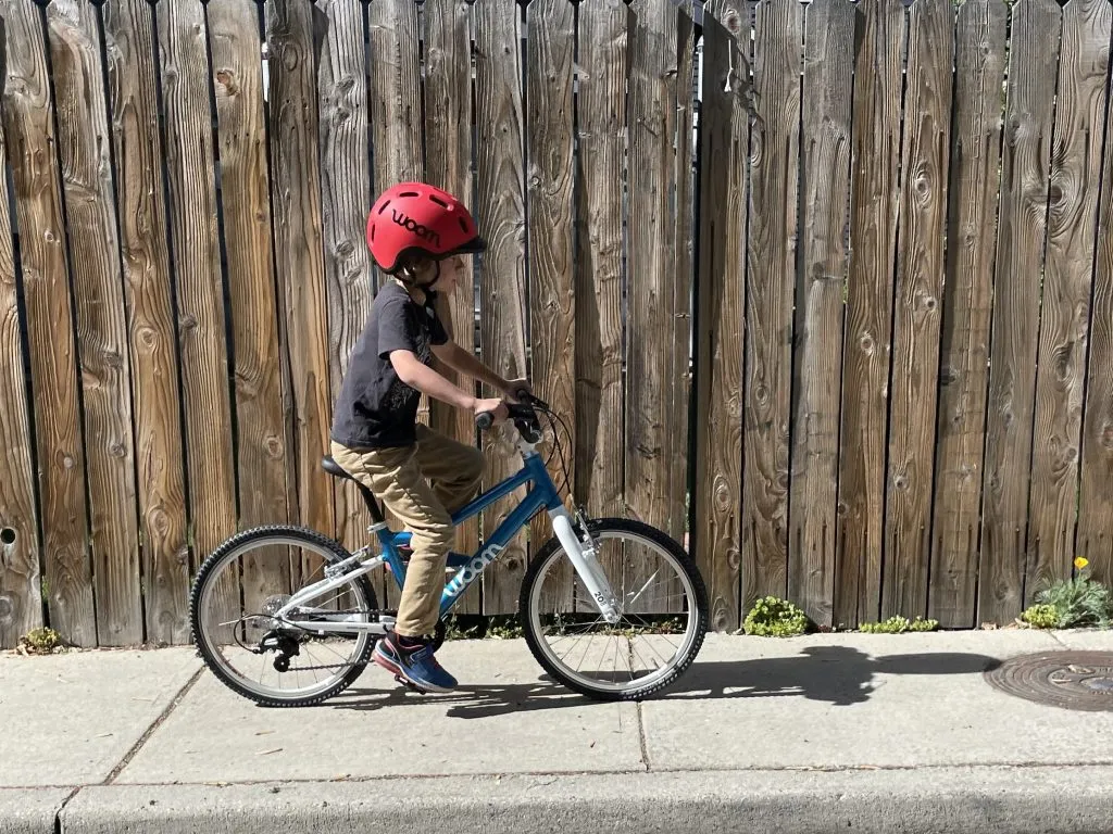 A child on a blue Woom bike with a white frame near a wooden fence