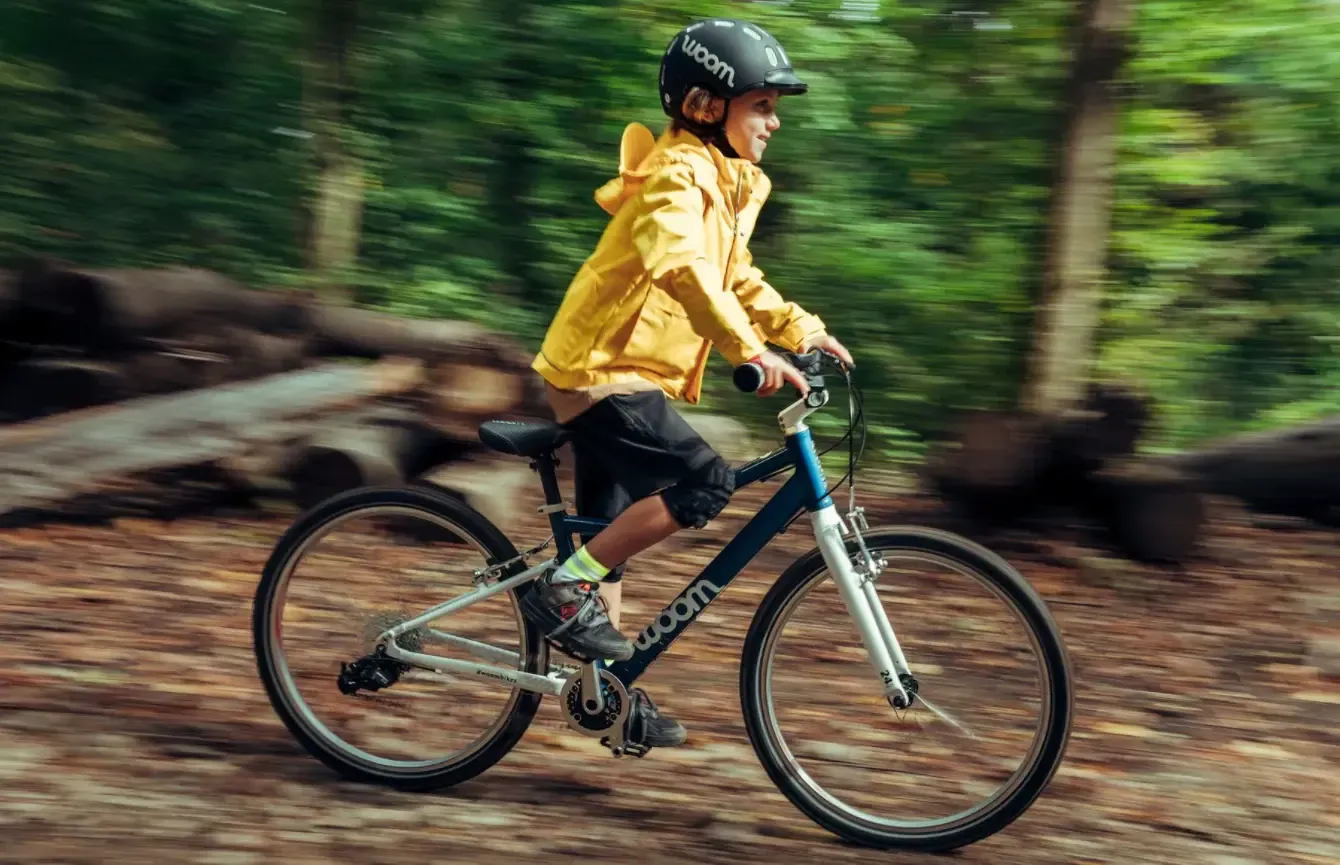 Woom children's bike in blue and silver, set against a forest backdrop, featuring a child wearing a black helmet