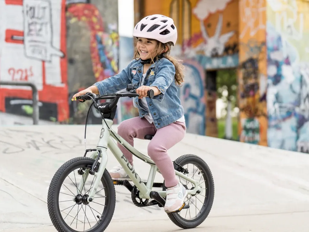 A smiling child on a light green Puky bike wearing a pink helmet in front of a colorful graffiti wall