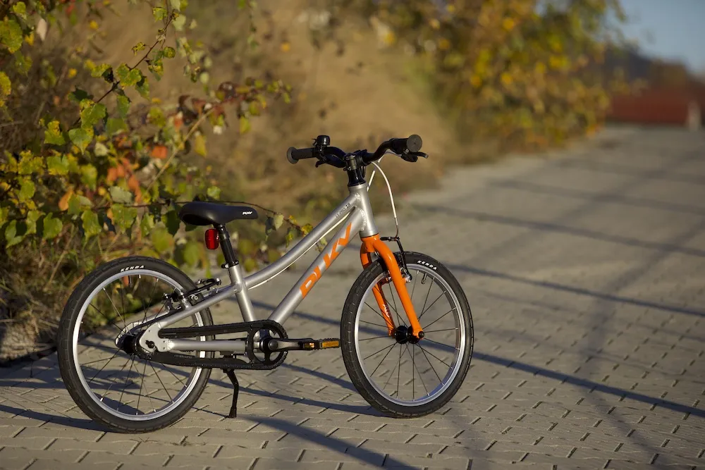 A silver and orange Puky children's bike on a sunny cobblestone path with foliage in the background
