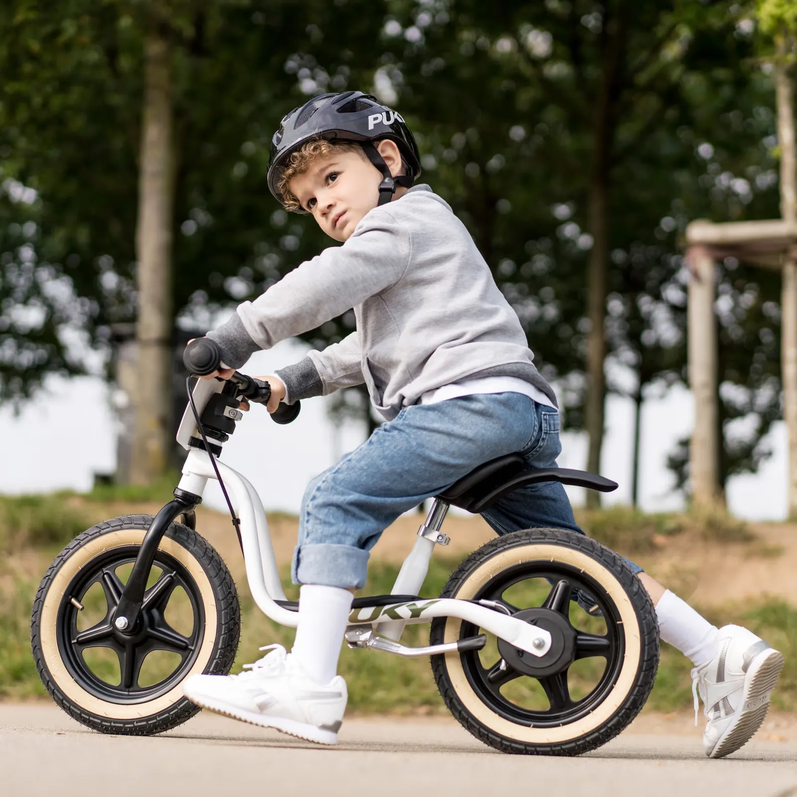 Child riding a white Puky balance bike outdoors, wearing a black helmet on a concrete path