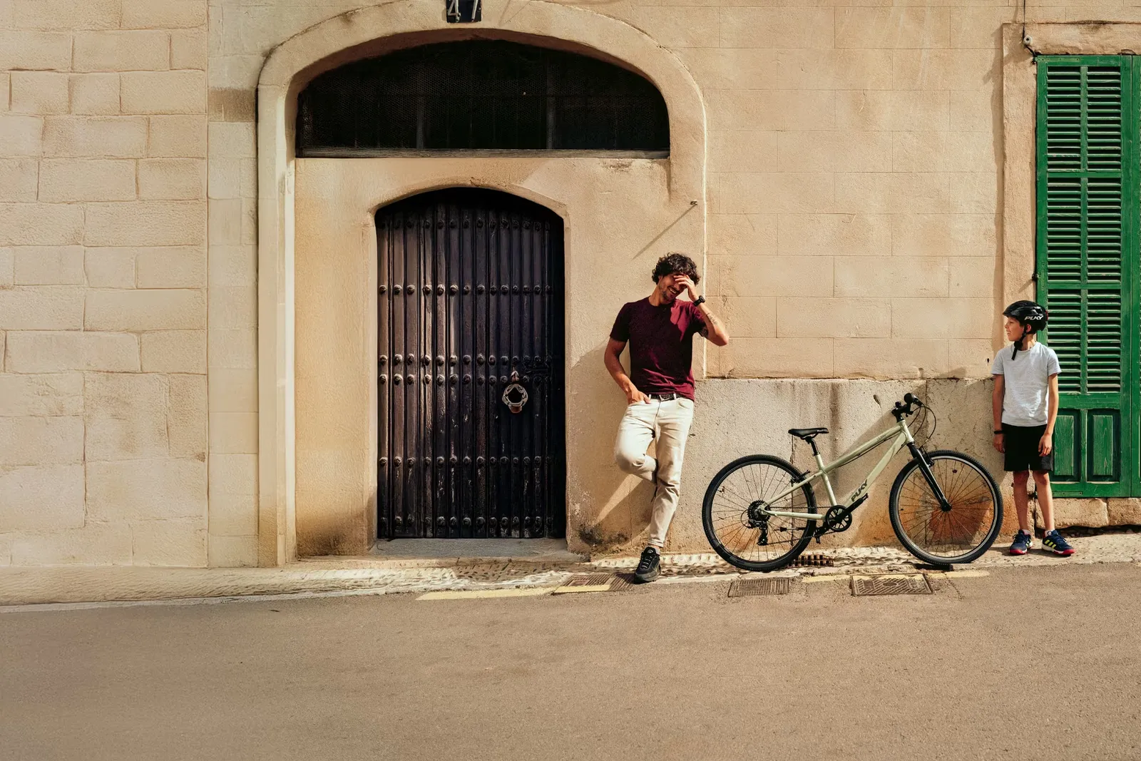 A beige Puky bike leaning against a beige wall, with a man and a child standing next to it, a black door, and a green window