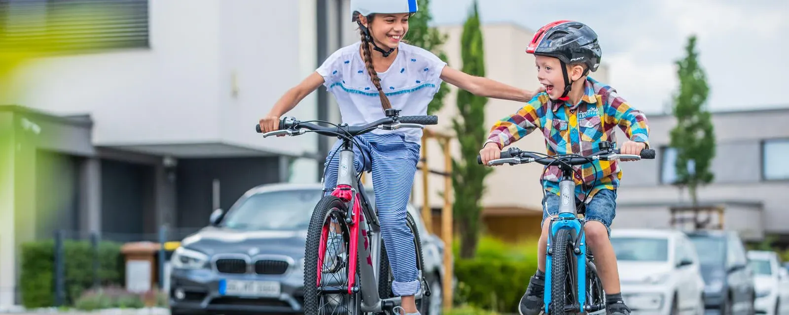 Two smiling children on colorful Puky bikes, wearing helmets, in a residential neighborhood