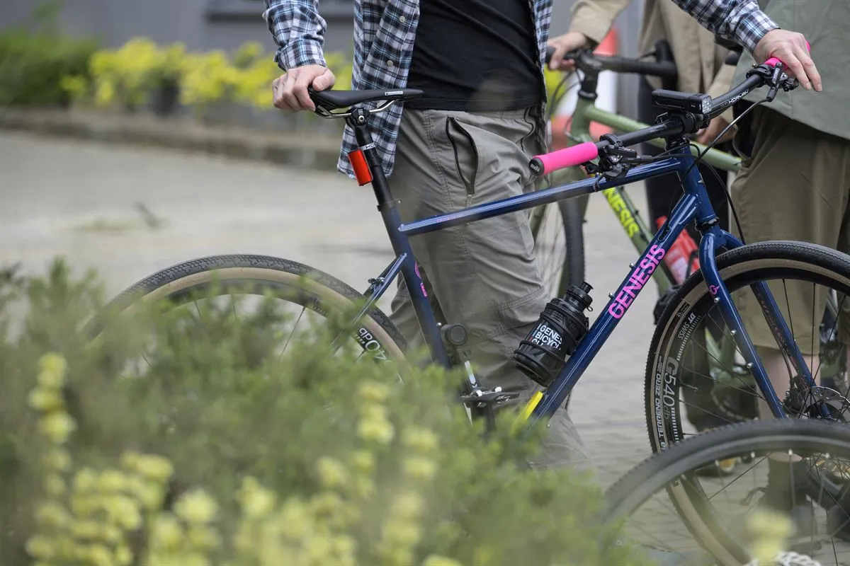 Vélo Genesis bleu foncé, cadre robuste, guidon rose, en extérieur urbain avec végétation en avant-plan