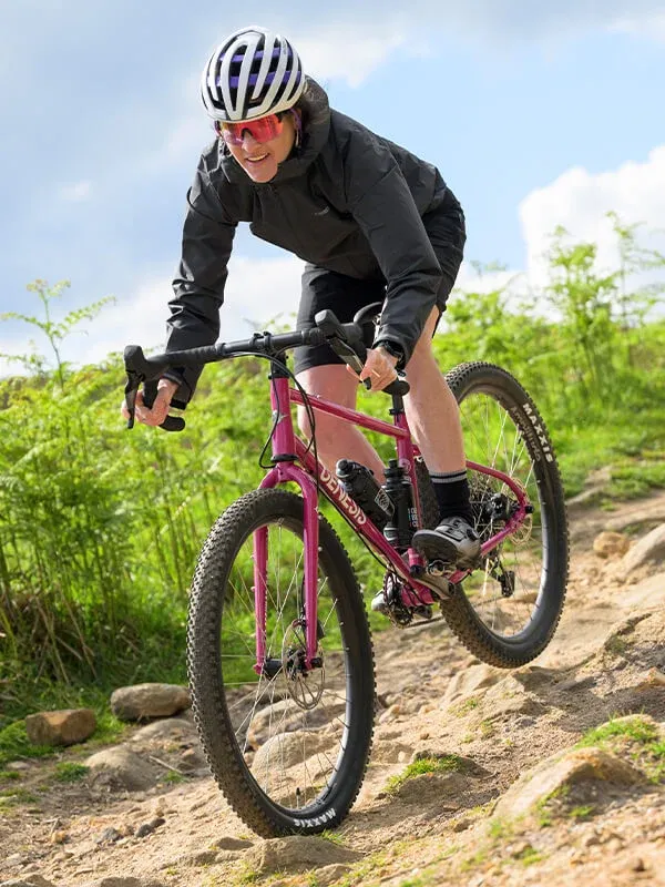 Vélo gravel Genesis rose sur sentier rocailleux, cycliste en tenue noire et casque rayé