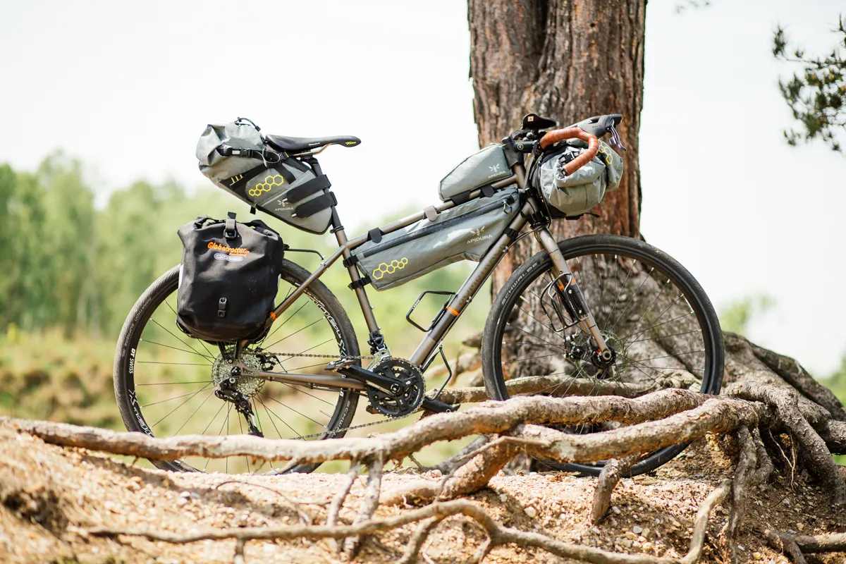 Gray Bombtrack touring bike with panniers and a sturdy frame, parked near a tree in a natural setting