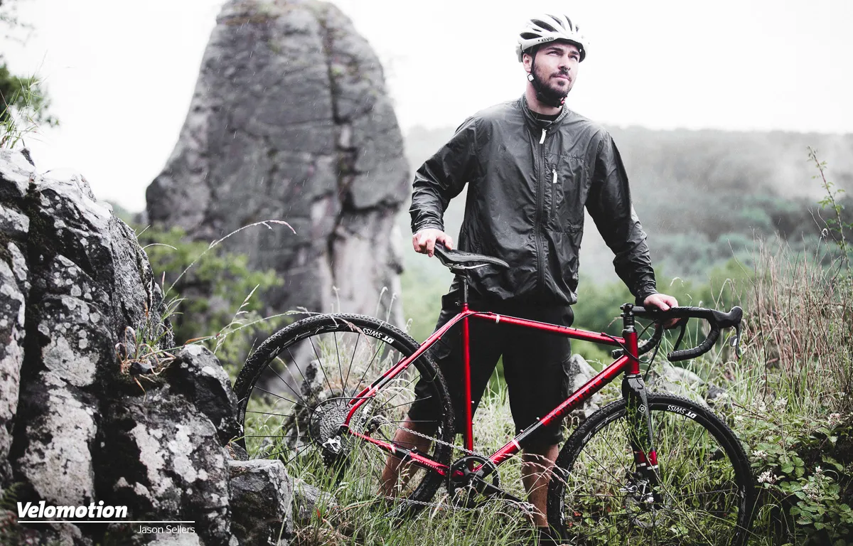 Red Bombtrack bike with a sturdy frame; a cyclist in outdoor gear on rocky, green terrain