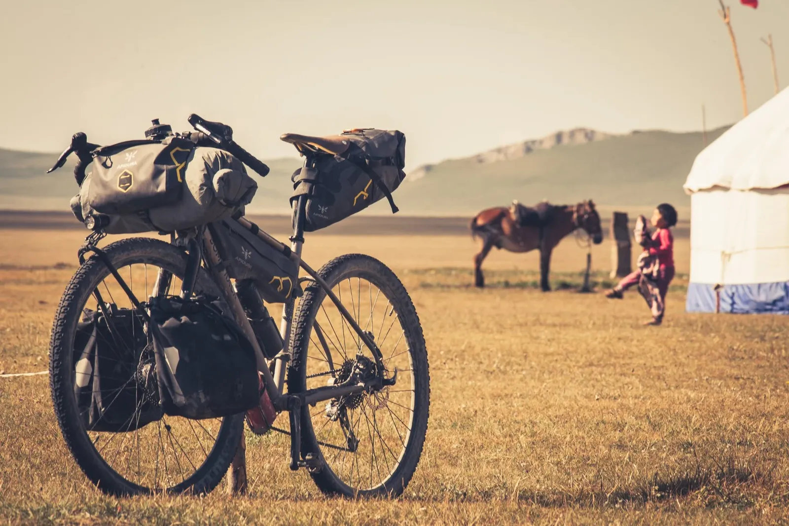 A Bombtrack bike with panniers on grassy terrain, with mountains and a yurt in the background