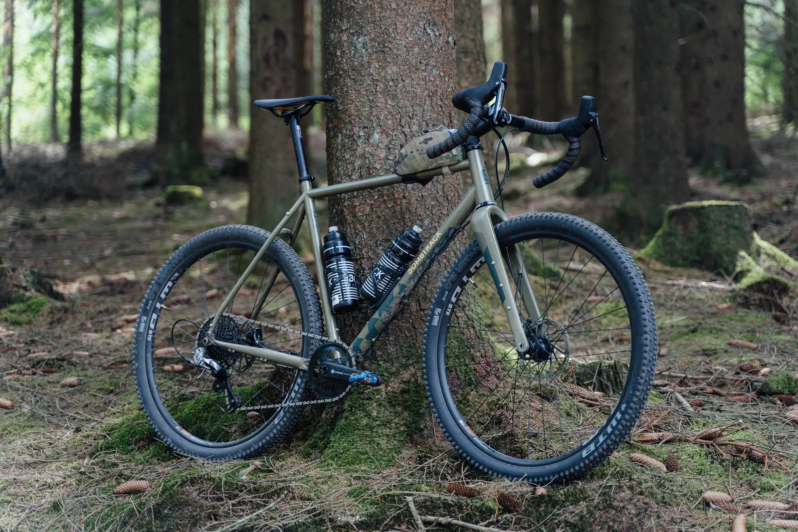 Khaki Bombtrack gravel bike with a sturdy frame, leaning against a tree in the forest