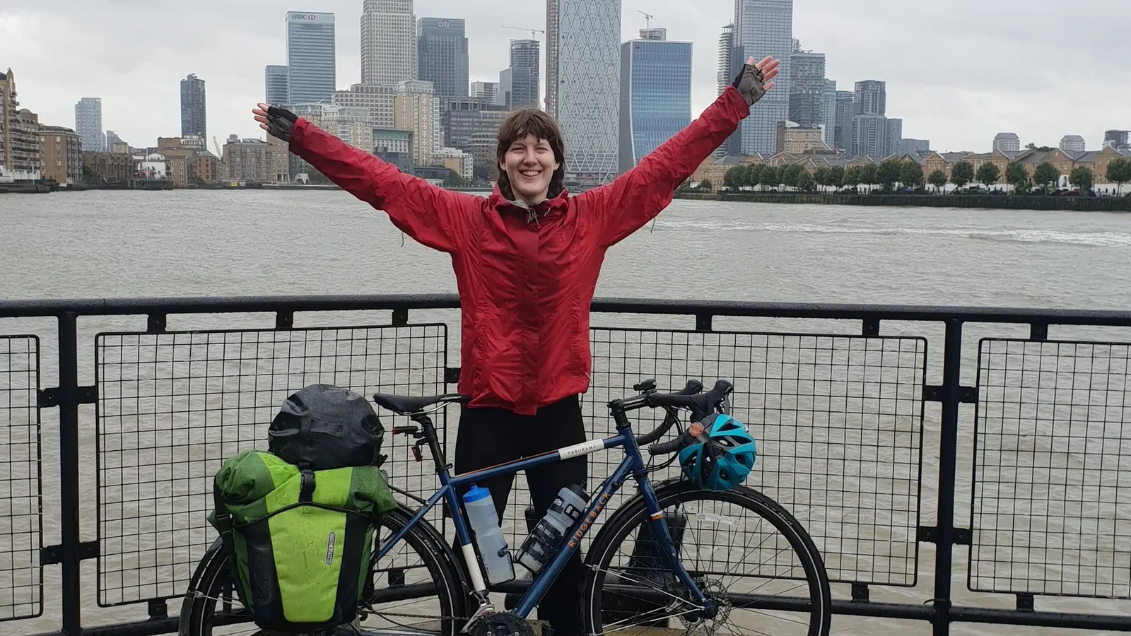 Blue Ridgeback bike with green panniers against a backdrop of an urban skyline and a river