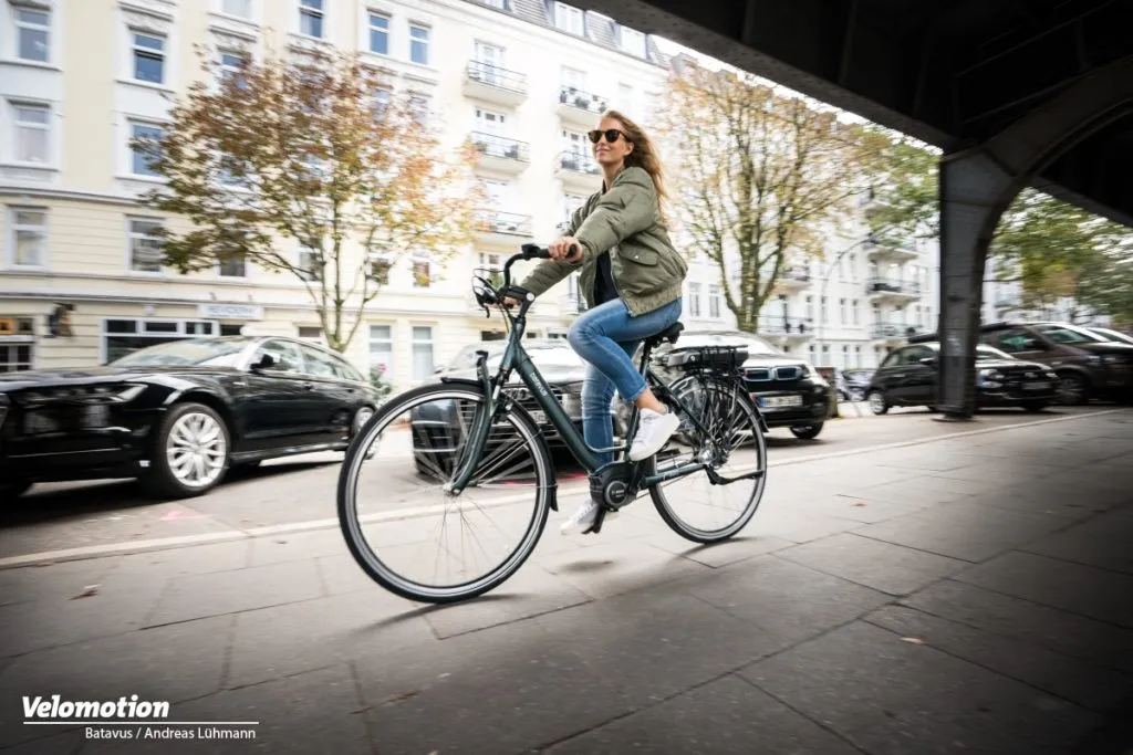 Black Batavus city bike, smiling woman riding under a bridge, stylish urban setting
