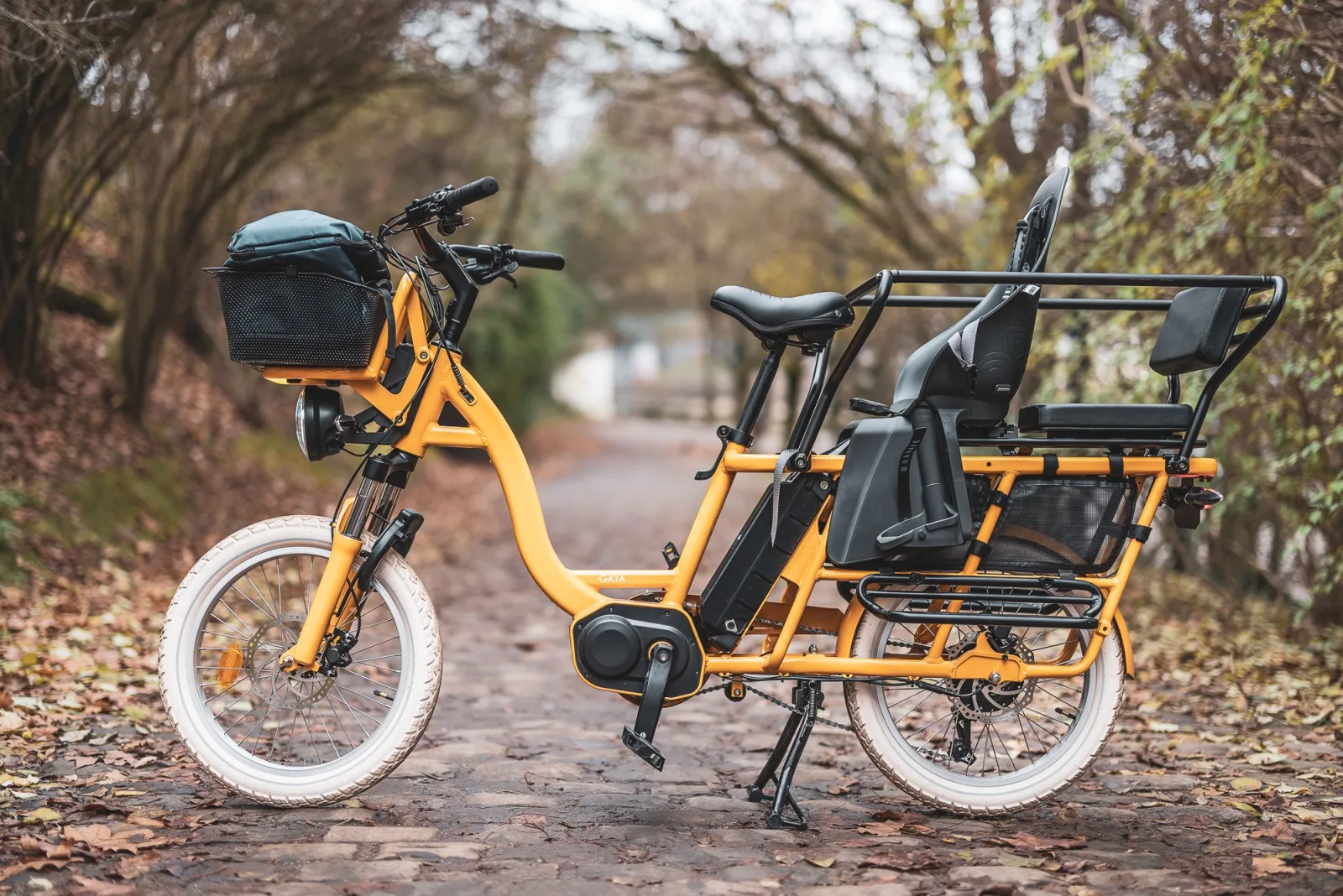 Vélo longtail électrique Gaya jaune avec siège enfant et panier, sur un chemin pavé en automne