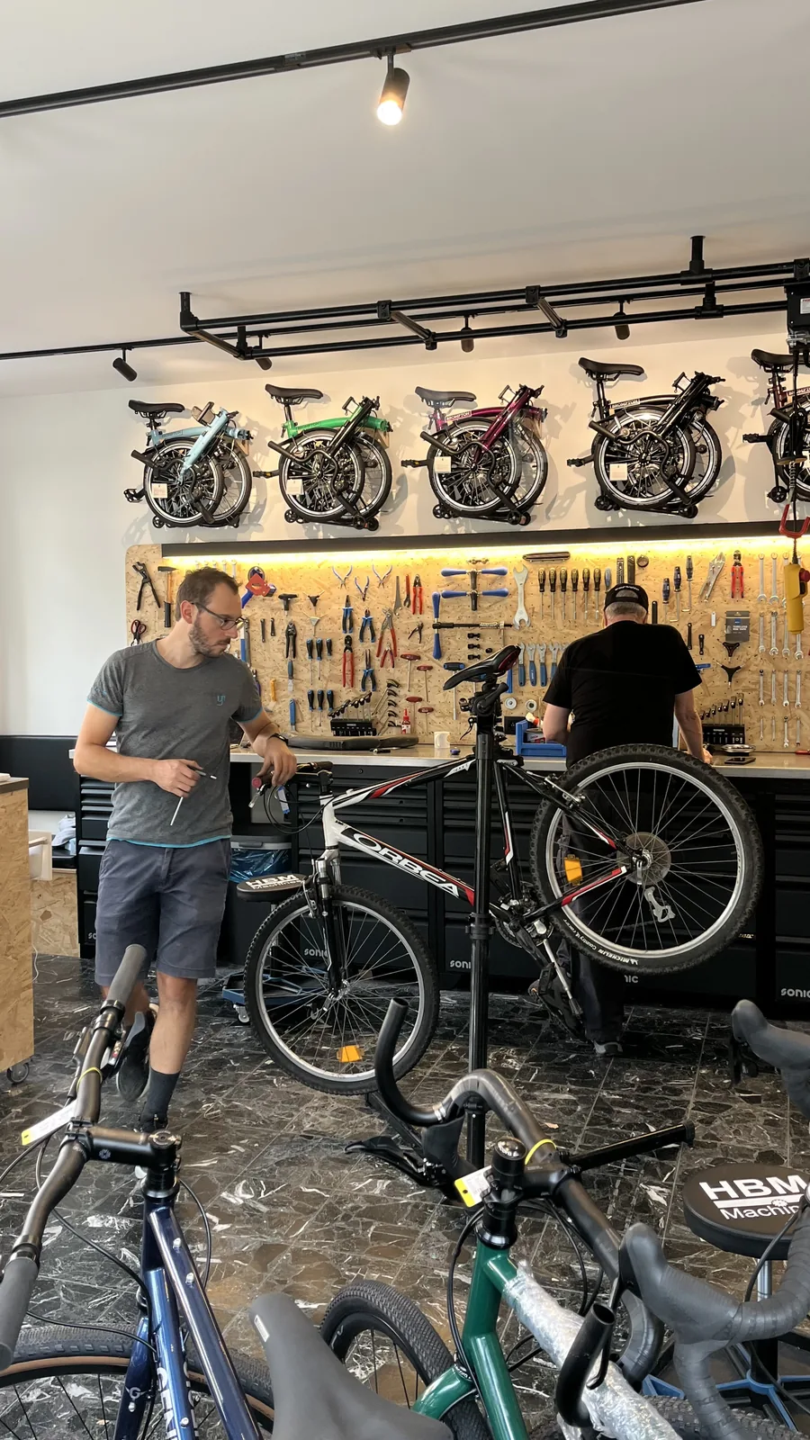 Deux employés dans un magasin de vélos s'affairent autour de plusieurs vélos et outils sur un mur d'atelier.