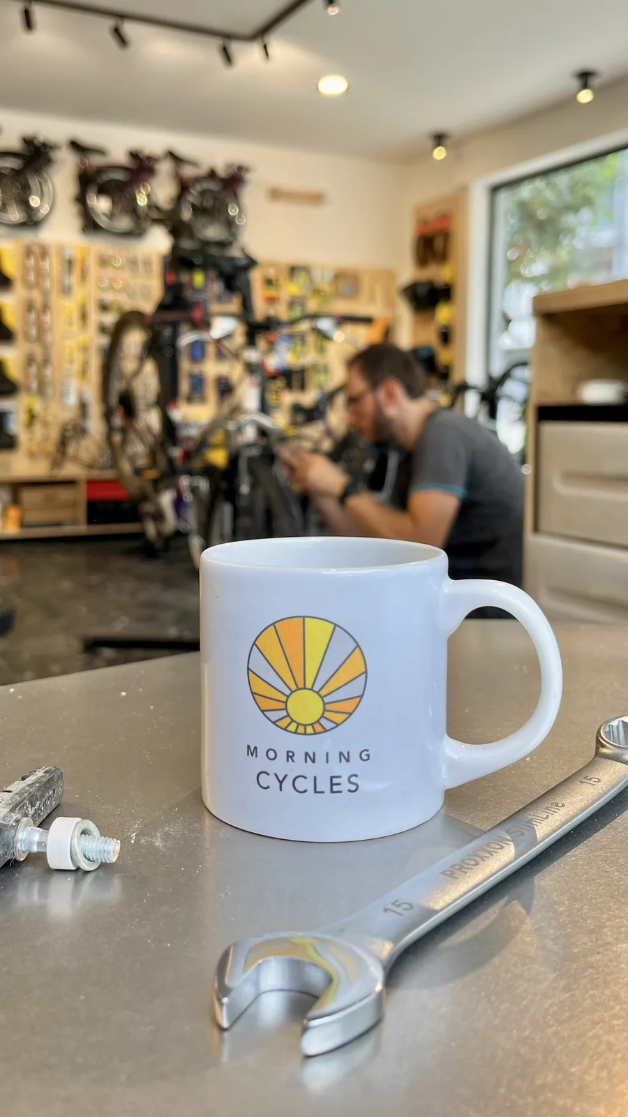 Tasse avec le logo Morning Cycles au premier plan, mécanicien travaillant sur un vélo en arrière-plan.