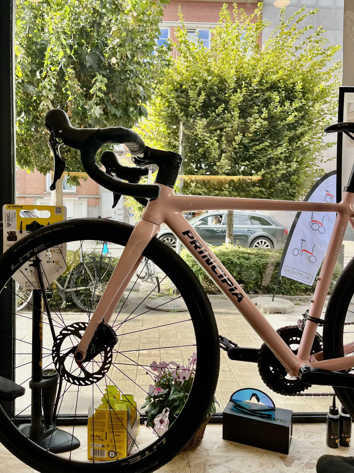 Un vélo de route rose est exposé derrière une vitrine, avec des arbres et des voitures visibles à l'extérieur.