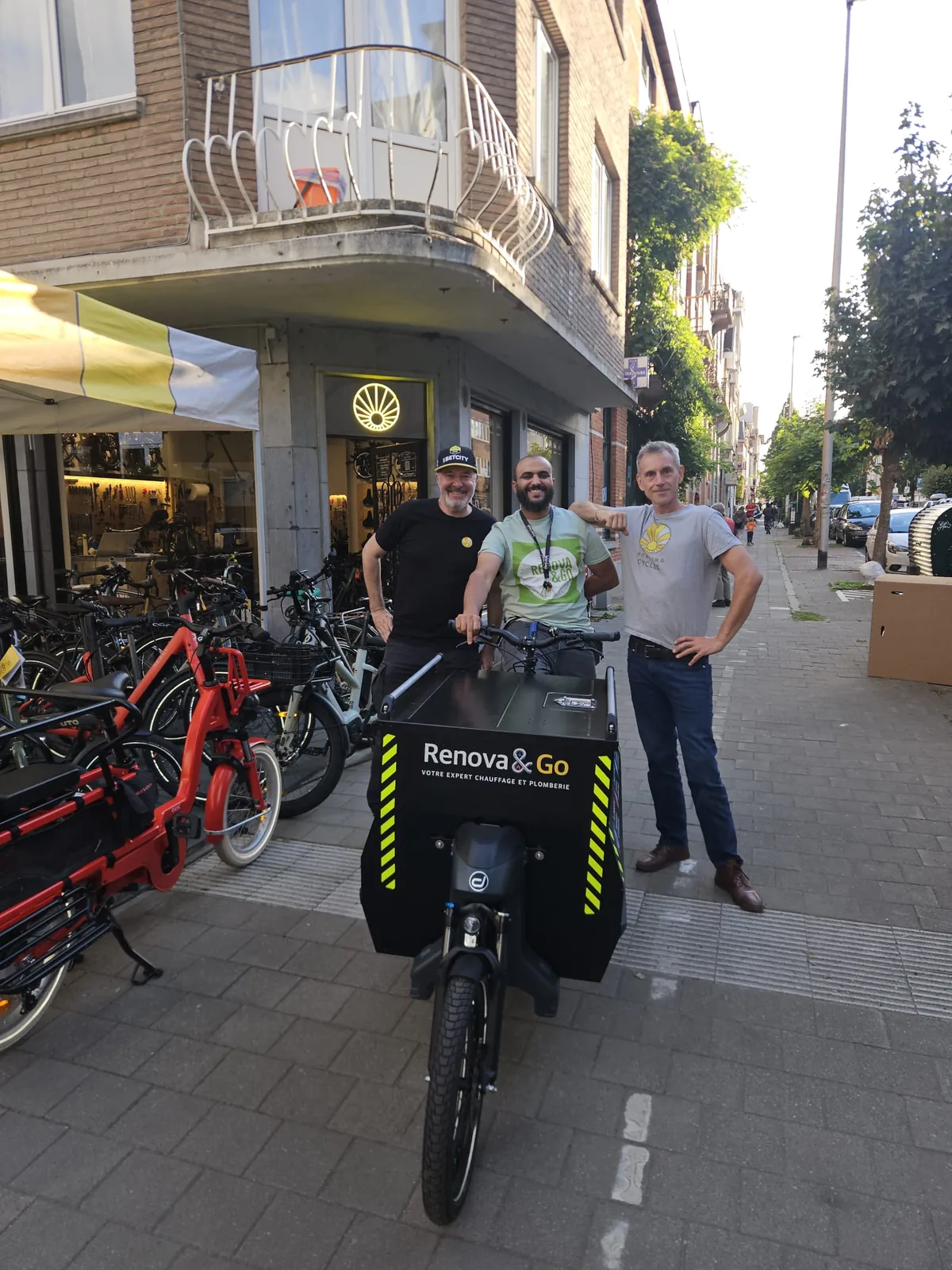 Trois personnes posent devant un vélo cargo avec l'inscription "Renova&Go" sur une rue pavée près d'un magasin de vélos.